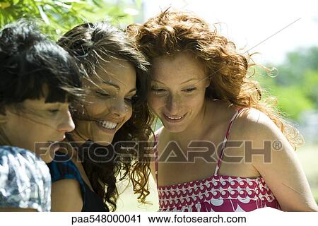 Stock Image - "Three young women outdoors, looking down, all smiling". Fotosearch