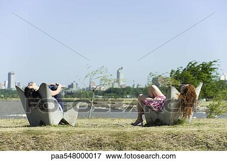 Stock Photo - "Two young women relaxing in chairs, pond and city skyline in background". Fotosearch