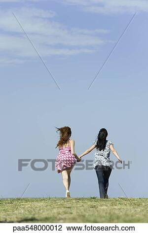 Stock Image - "Two young women running hand in hand, rear view". Fotosearch