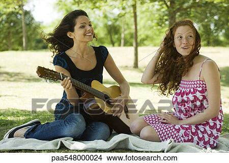 "Two young women sitting on blanket outdoors, one playing acoustic guitar" View Large Photo Image Picture - "Two young women sitting on blanket outdoors, one playing acoustic guitar". Fotosearch