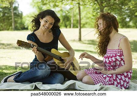 "Two young women sitting on blanket outdoors, one playing acoustic guitar" View Large Photo Image Stock Photograph - "Two young women sitting on blanket outdoors, one playing acoustic guitar". Fotosearch
