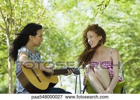"Two young women sitting outdoors, one playing acoustic guitar" View Large Photo Image Stock Photo - "Two young women sitting outdoors, one playing acoustic guitar". Fotosearch