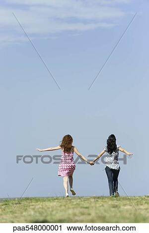 Stock Image - "Two young women walking hand in hand outdoors, rear view". Fotosearch