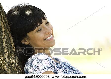 "Young woman leaning against tree trunk, smiling" View Large Photo Image Stock Photo - "Young woman leaning against tree trunk, smiling". Fotosearch