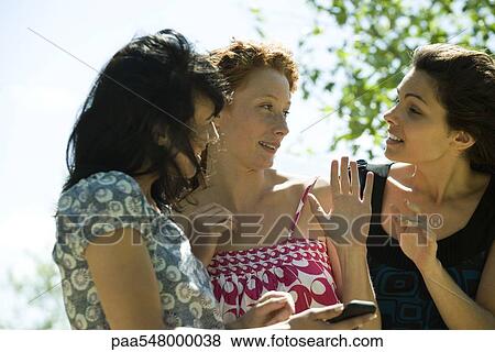 "Young women chatting outdoors, one holding cell phone" View Large Photo Image Stock Photo - "Young women chatting outdoors, one holding cell phone". Fotosearch