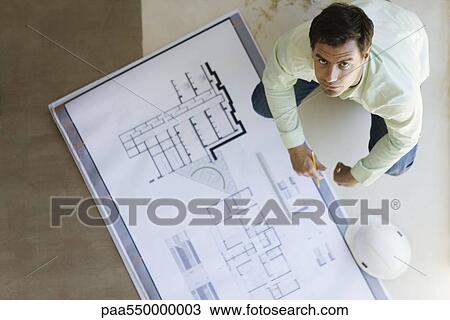 Stock Image - "Architect crouching looking up at camera, blueprints spread out on floor". Fotosearch