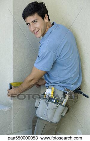 Stock Image - "Construction worker measuring space on wall, looking over shoulder at camera". Fotosearch