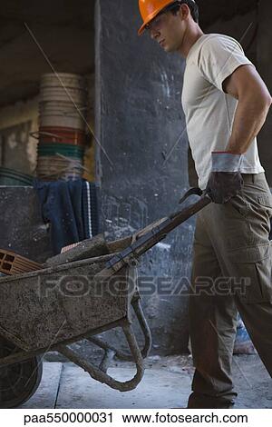 Stock Image - Construction worker pushing wheelbarrow through construction site. Fotosearch