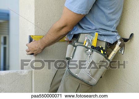 Stock Image - Construction worker wearing tool belt heavy with tools measuring wall. Fotosearch