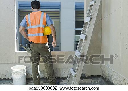 Stock Image - "Construction worker with hand on hip looking out window, rear view". Fotosearch