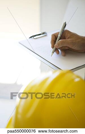 Stock Photograph - "Hand writing notes on clipboard pad, hard hat in foreground, cropped view". Fotosearch