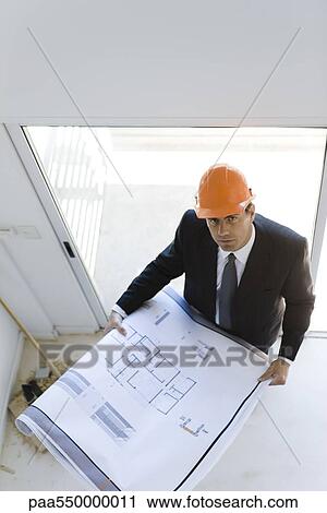 Stock Image - Man in suit wearing hard hat holding blueprints looking up at camera. Fotosearch
