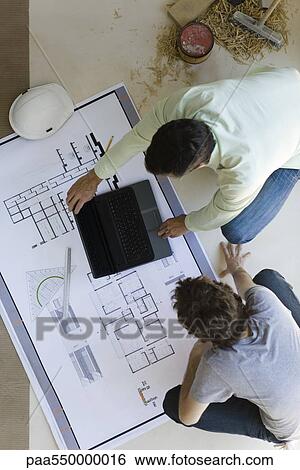 Stock Photograph - "Two men crouching looking over blueprints, using laptop set on floor". Fotosearch