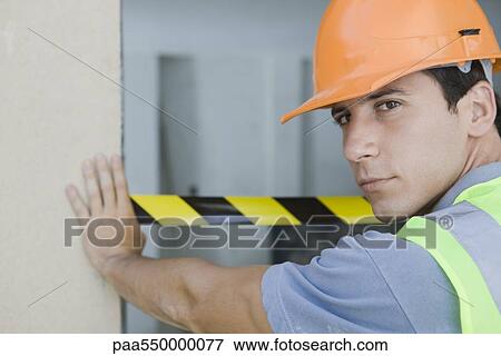 Stock Photo - "Worker placing safety tape across doorway, looking over shoulder at camera". Fotosearch