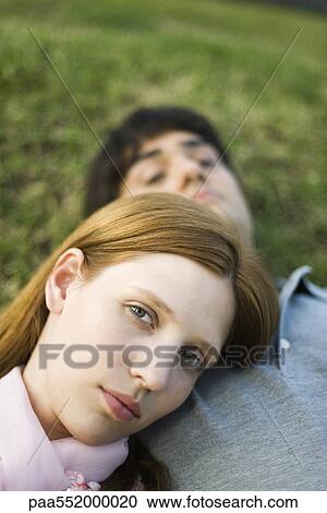 Stock Image - "Young couple lying on ground, woman resting head on man's chest, looking at camera". Fotosearch