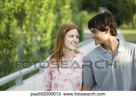 Stock Image - "Young couple walking together outdoors, smiling at each other". Fotosearch
