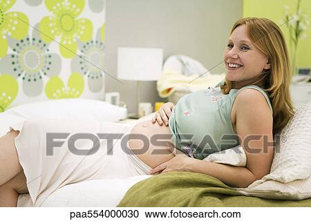 Stock Image - Pregnant woman relaxing on bed, smiling. Fotosearch