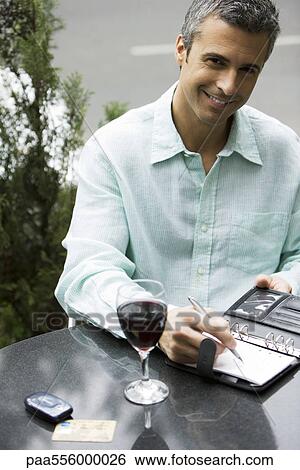 Man sitting at outdoor cafe table with glass of wine, writing in agenda View Large Photo Image Stock Photograph - Man sitting at outdoor cafe table with glass of wine, writing in agenda. Fotosearch