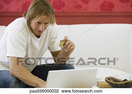 Stock Image - Man sitting on sofa, making online purchase with credit card. Fotosearch