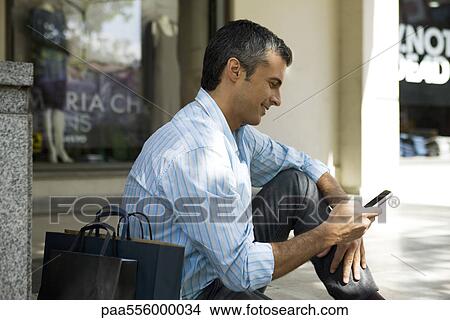 Picture - Man sitting with outdoors with shopping bags, using cell phone. Fotosearch