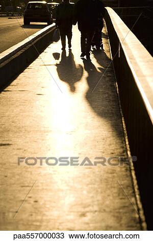 Stock Image - Pedestrians walking on sidewalk, backlit. Fotosearch