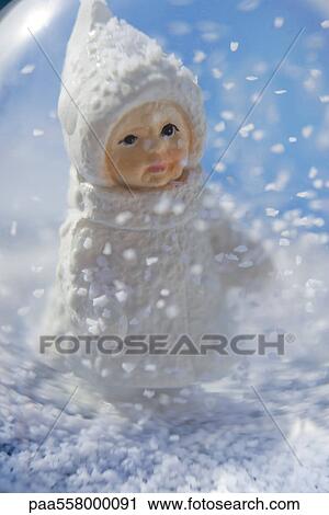 Stock Image - Snow globe, figurine of little girl wearing winter coat and hat looking at snow flakes falling. Fotosearch