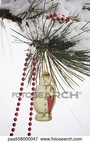 Stock Photo - Snowman Christmas ornament and beaded garland hanging from snow-covered tree branch. Fotosearch