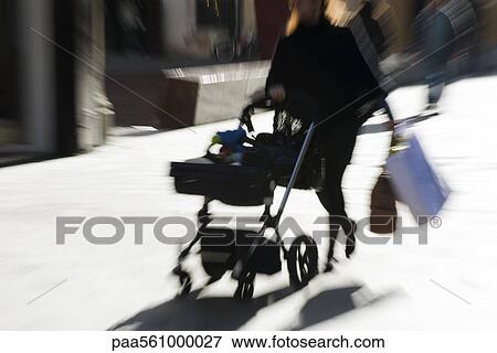 Stock Photo - Pedestrian pushing baby carriage on sidewalk, carrying shopping bags, blurred. Fotosearch