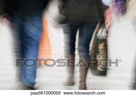 Stock Image - Pedestrians carrying shopping bags, rear view, blurred. Fotosearch