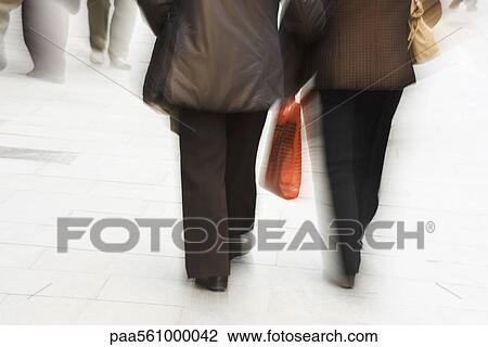 Stock Image - Pedestrians walking on sidewalk, one carrying shopping bag, rear view. Fotosearch