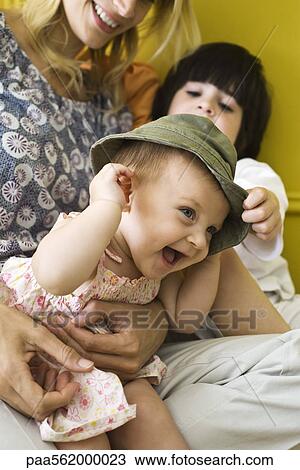 Stock Image - Infant girl sitting with mother and brother, wearing hat and laughing. Fotosearch
