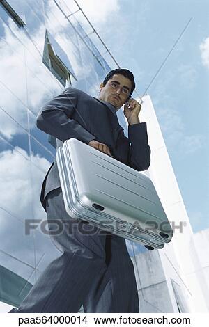 Picture - Businessman carrying briefcase using cell phone outside office building. Fotosearch