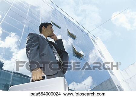 Picture - Businessman carrying briefcase using cell phone outside office building. Fotosearch