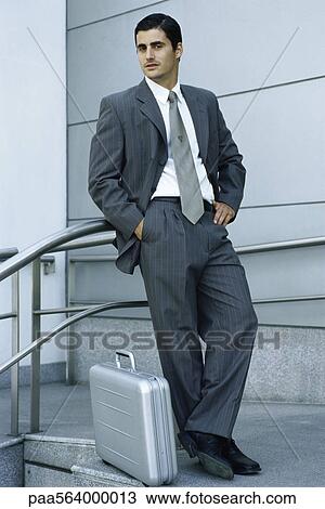 Stock Image - Businessman leaning against railing, briefcase on sidewalk at feet. Fotosearch