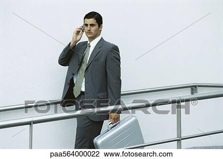 Stock Image - Businessman walking on sidewalk using cell phone, carrying briefcase. Fotosearch