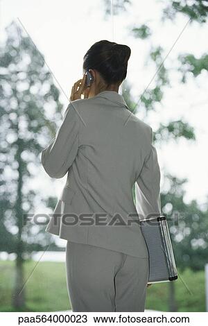 Stock Image - Businesswoman using cell phone, carrying briefcase under arm, rear view. Fotosearch