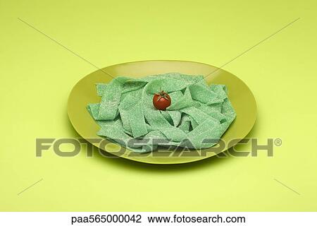 Stock Image - Food concept, plate of green gummy candy arranged like pasta topped with cherry tomato. Fotosearch