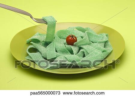 Stock Image - Food concept, plate of green gummy candy arranged like pasta, topped with cherry tomato. Fotosearch