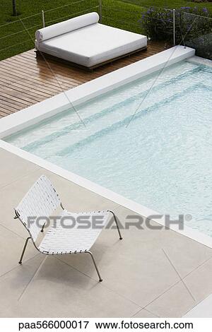 Stock Photo - Chair at edge of pool, sunbathing bed on deck in background. Fotosearch