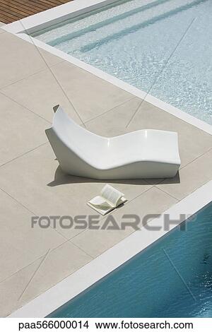 Picture - Open book lying on ground next to poolside deckchair, high angle view. Fotosearch
