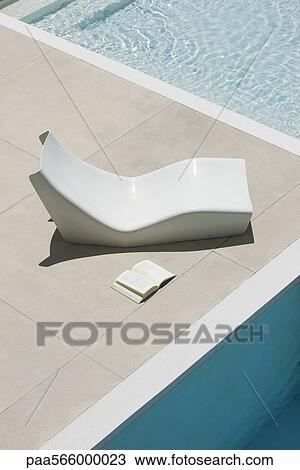 Stock Image - Open book lying on ground next to poolside deckchair. Fotosearch