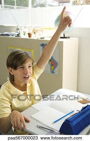 Stock Image - Boy sitting in classroom, raising hand enthusiastically. Fotosearch