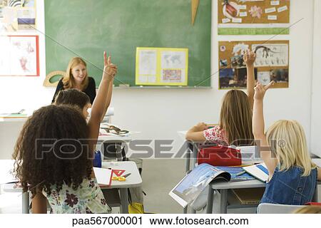 Stock Image - Elementary students in classroom raising hands, rear view. Fotosearch