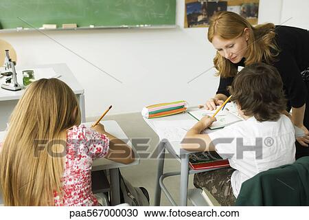 Elementary Teacher Leaning Over Student S Desk Helping Him With