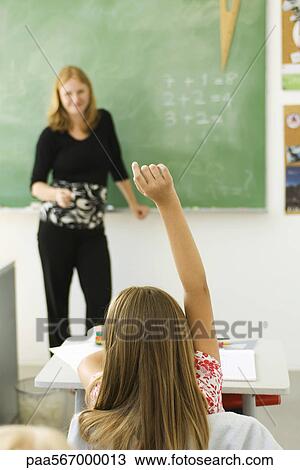 Stock Image - Girl in classroom raising hand to answer question. Fotosearch