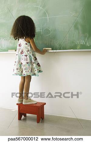 Stock Image - Little girl standing on stool in front of blackboard, rear view. Fotosearch