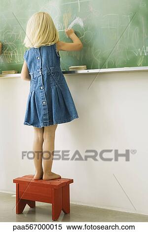 Stock Photography - Little girl standing on stool, scribbling on blackboard, rear view. Fotosearch
