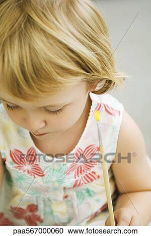 Stock Image - Toddler girl holding paintbrush, looking down. Fotosearch