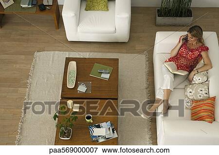 Stock Photo - Woman relaxing on sofa with book on lap, talking on cell phone. Fotosearch