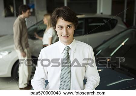 Stock Image - Car salesman in car dealership showroom, potential buyers in background. Fotosearch